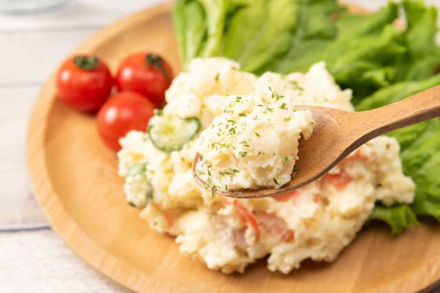 木製の皿に盛られたきゅうりやにんじん入りの冷製ポテトサラダ。木のスプーンですくい、レタスとミニトマトを添えた写真